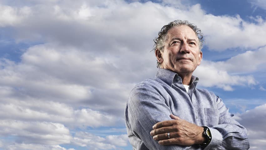Tom Siebel, founder and CEO of C3 AI, standing with his arms crossed and looking into the distance, against a backdrop of blue sky and white clouds.
